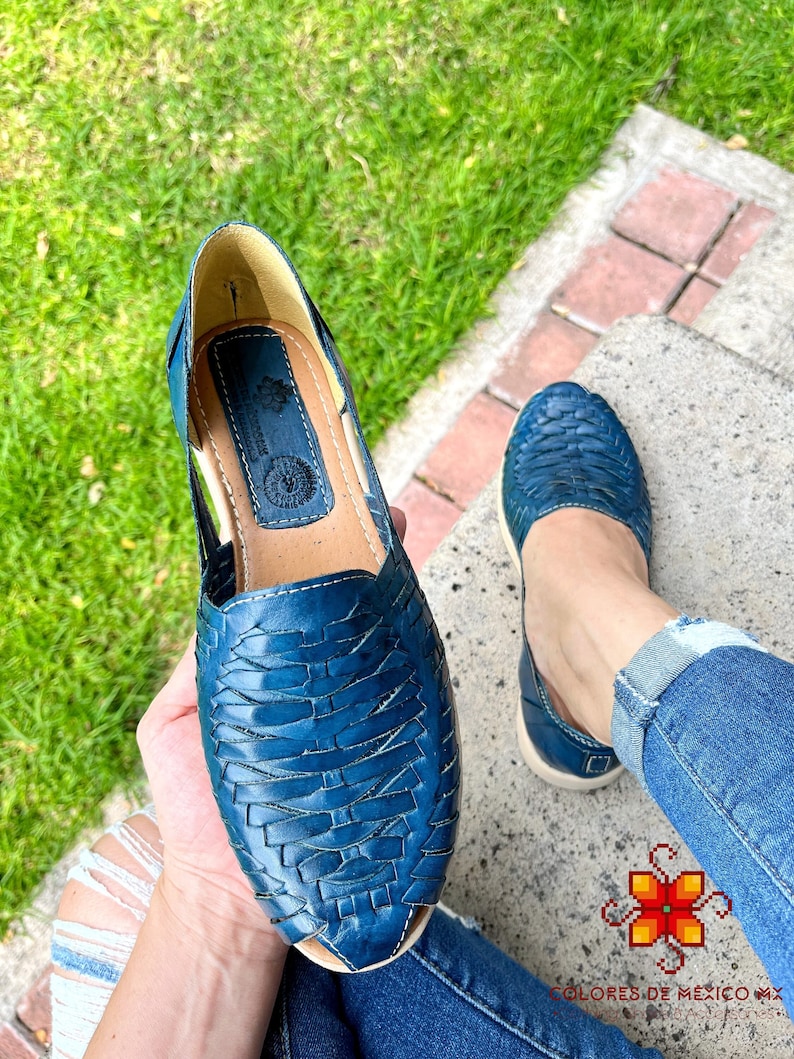May include: A pair of blue leather sandals with a woven design. The sandals have a pointed toe and an open back. The sandals are being held by a person's hand.