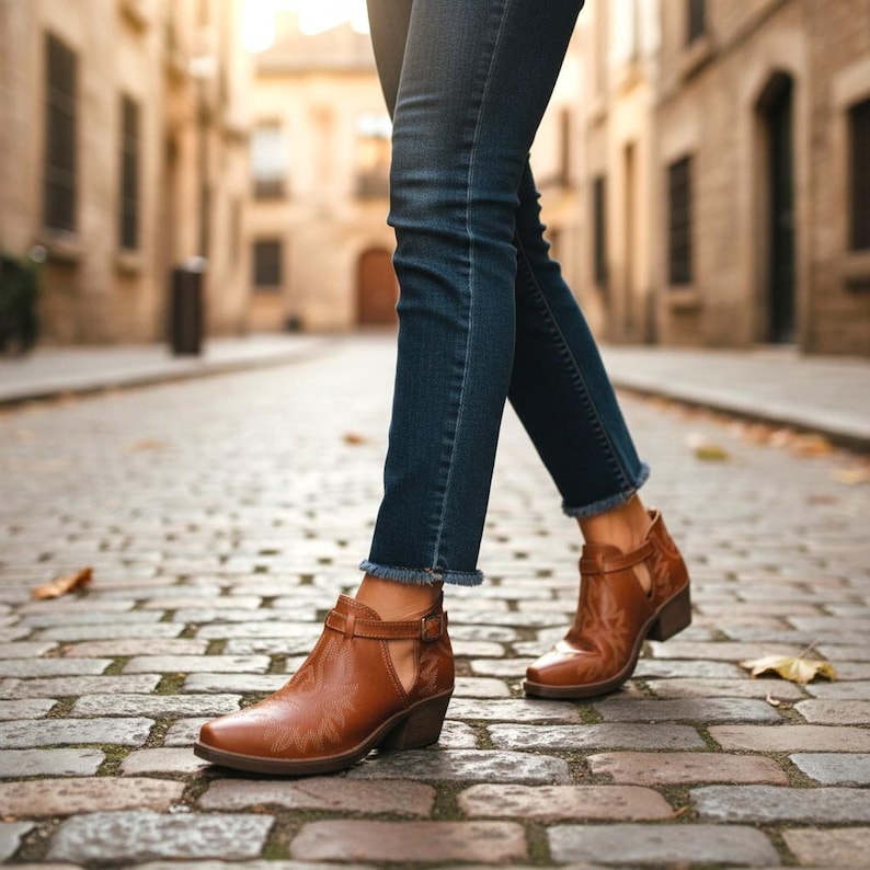 Pu&ograve; includere: Stivaletti in pelle marrone con un design cucito decorativo e un piccolo tacco. Gli stivaletti hanno un dettaglio laterale ritagliato e una chiusura con fibbia. Gli stivaletti sono indossati con jeans blu.