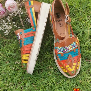 May include: A pair of colorful leather huaraches sandals with a woven design in shades of orange, yellow, blue, and green. The sandals have a brown leather upper and a white sole. The sandals are sitting on green grass.