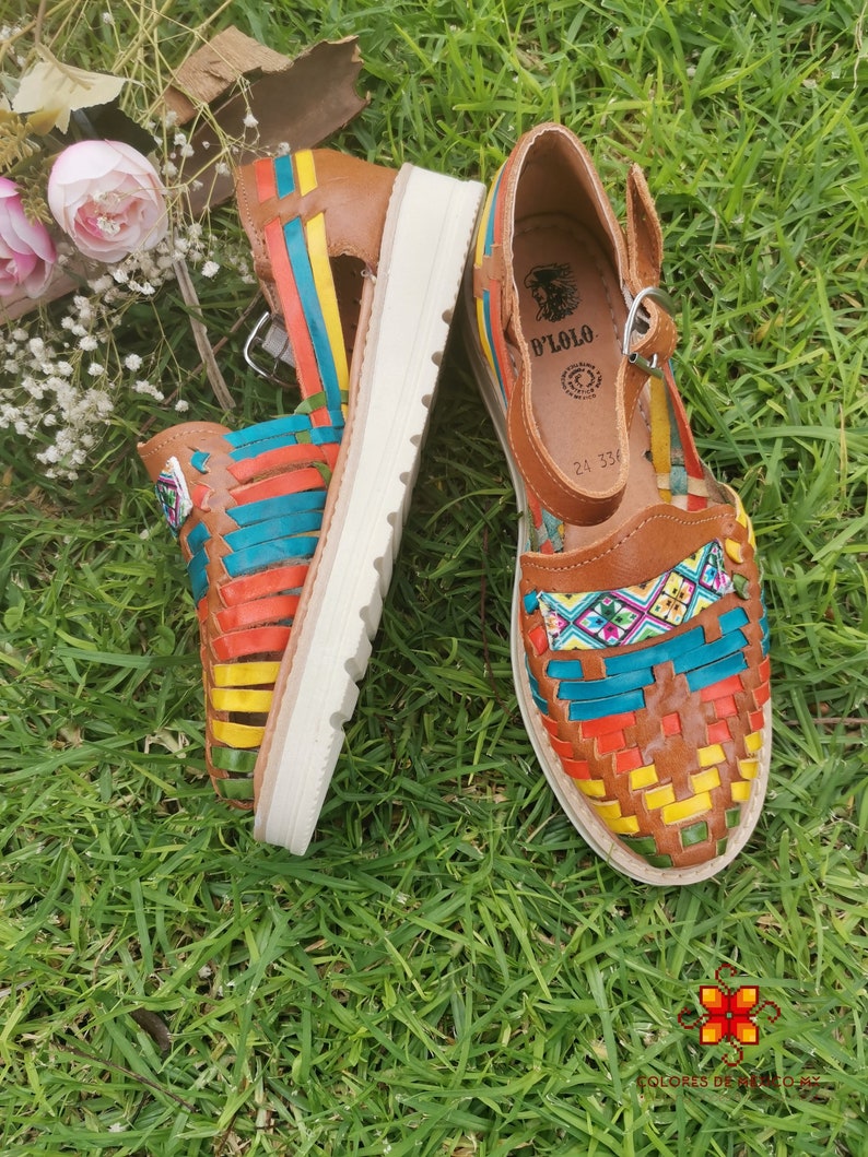 May include: A pair of colorful leather huaraches sandals with a woven design in shades of orange, yellow, blue, and green. The sandals have a brown leather upper and a white sole. The sandals are sitting on green grass.