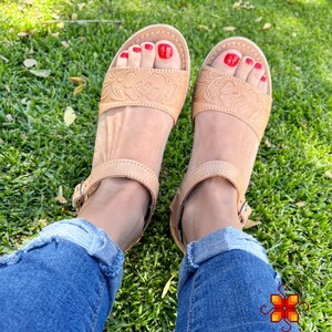 Peut inclure: Sandales en cuir tann&eacute; avec des motifs floraux complexes, dot&eacute;es d'une fermeture &agrave; boucle et d'un vernis &agrave; ongles rouge sur les orteils. Les sandales sont port&eacute;es par une personne v&ecirc;tue d'un jean bleu et assise sur de l'herbe verte.