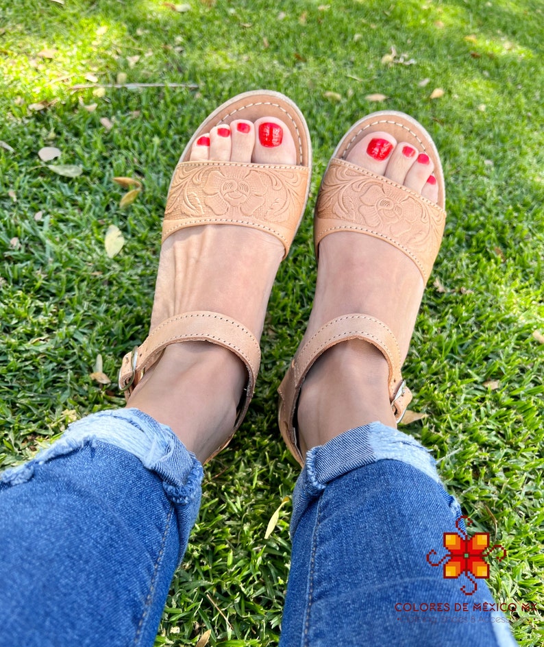 Peut inclure: Sandales en cuir tann&eacute; avec des motifs floraux complexes, dot&eacute;es d'une fermeture &agrave; boucle et d'un vernis &agrave; ongles rouge sur les orteils. Les sandales sont port&eacute;es par une personne v&ecirc;tue d'un jean bleu et assise sur de l'herbe verte.
