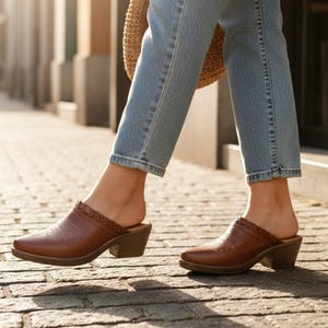 May include: Brown leather mules with a pointed toe and a low block heel. The shoes feature a decorative stitched design on the vamp and a braided trim around the opening. The person is wearing light blue jeans.