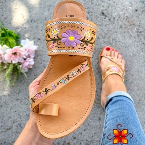 K&ouml;nnte beinhalten: Tanfarbene Ledersandalen mit floralem Stickmuster. Die Sandalen haben eine Zehenschlaufe und einen Riemen, der um den Kn&ouml;chel geht. Die Stickerei zeigt rosa, lila, gelbe und gr&uuml;ne Blumen.