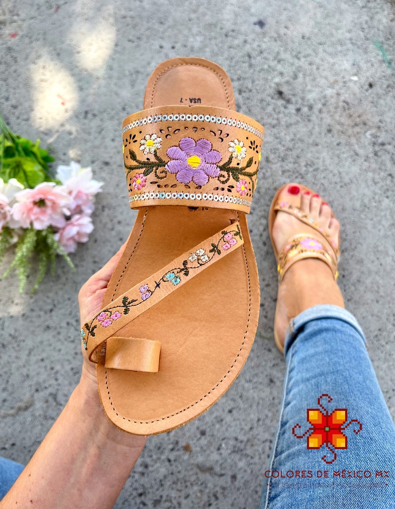 K&ouml;nnte beinhalten: Tanfarbene Ledersandalen mit floralem Stickmuster. Die Sandalen haben eine Zehenschlaufe und einen Riemen, der um den Kn&ouml;chel geht. Die Stickerei zeigt rosa, lila, gelbe und gr&uuml;ne Blumen.