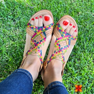 May include: A pair of woven leather sandals with a multi-coloured design. The sandals have a criss-cross strap design and are being worn on a person's feet. The person is wearing blue jeans.