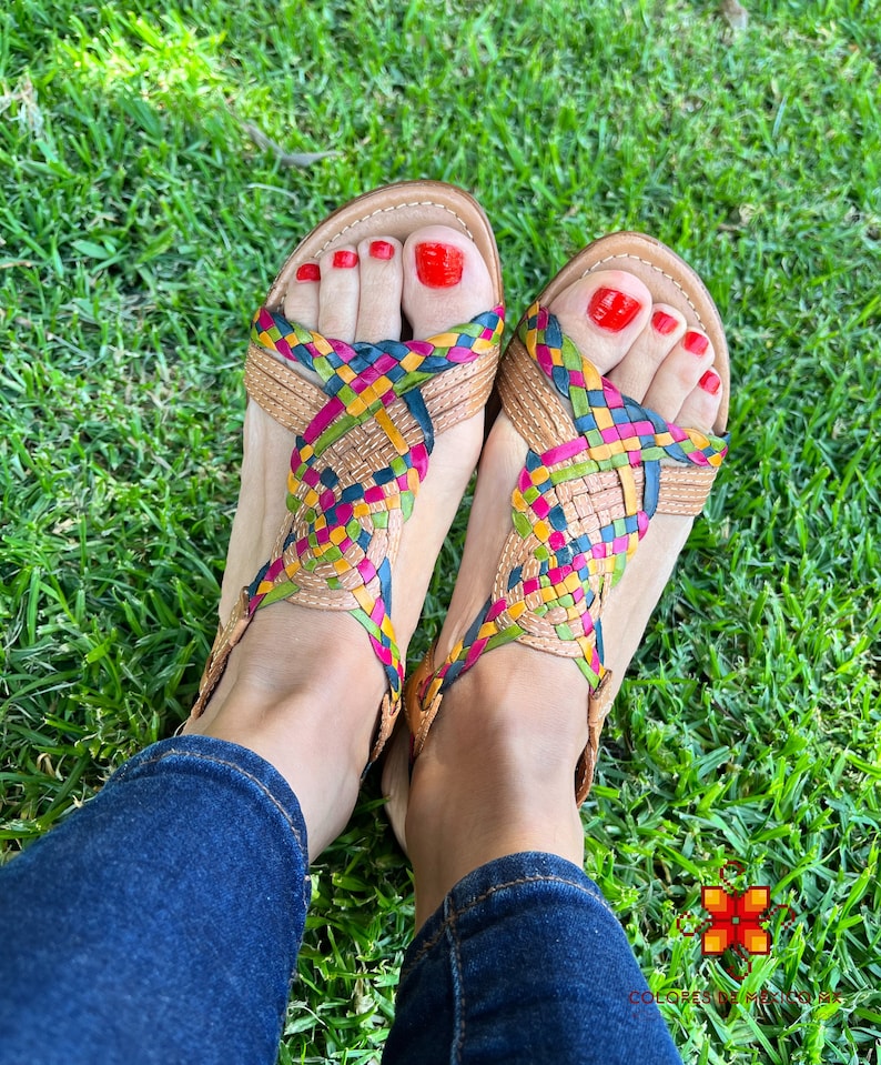 May include: A pair of woven leather sandals with a multi-coloured design. The sandals have a criss-cross strap design and are being worn on a person's feet. The person is wearing blue jeans.