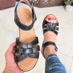 May include: Black leather sandals with a woven design and an adjustable ankle strap. The sandals have a brown insole and a wooden platform sole. The brand name "Cucos" is visible on the insole.