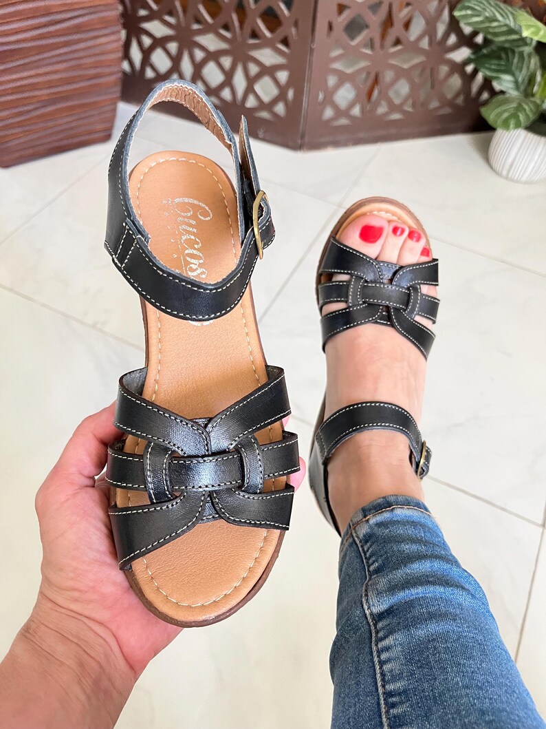 May include: Black leather sandals with a woven design and an adjustable ankle strap. The sandals have a brown insole and a wooden platform sole. The brand name "Cucos" is visible on the insole.