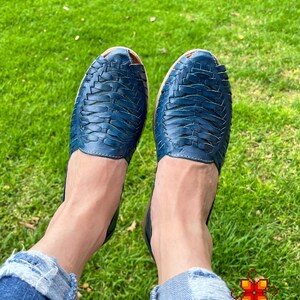May include: A pair of blue leather sandals with a woven design. The sandals are being worn by someone sitting on a grassy surface.