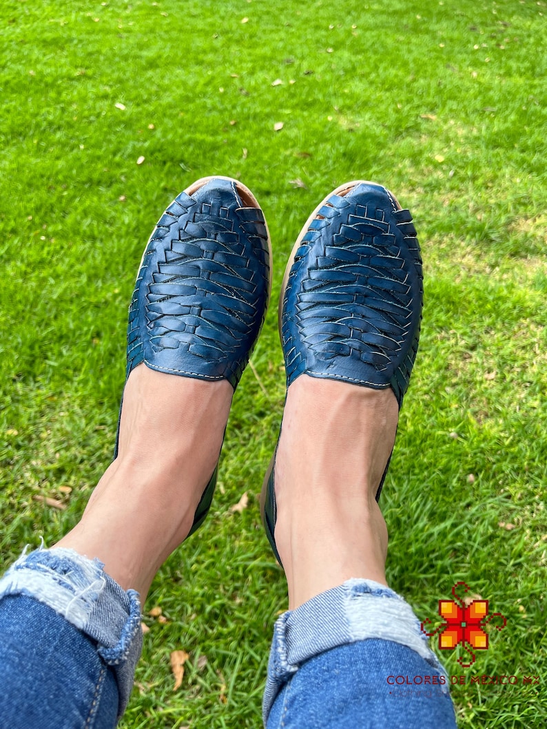 May include: A pair of blue leather sandals with a woven design. The sandals are being worn by someone sitting on a grassy surface.
