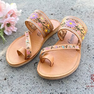 K&ouml;nnte beinhalten: Ein Paar braune Ledersandalen mit Blumenstickerei. Die Sandalen haben eine Zehenschlaufe und zwei Riemen, die um den Kn&ouml;chel gehen. Die Stickerei ist in den Farben Rosa, Lila, Gr&uuml;n und Gelb gehalten.