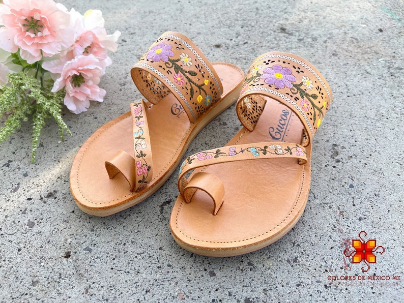 K&ouml;nnte beinhalten: Ein Paar braune Ledersandalen mit Blumenstickerei. Die Sandalen haben eine Zehenschlaufe und zwei Riemen, die um den Kn&ouml;chel gehen. Die Stickerei ist in den Farben Rosa, Lila, Gr&uuml;n und Gelb gehalten.