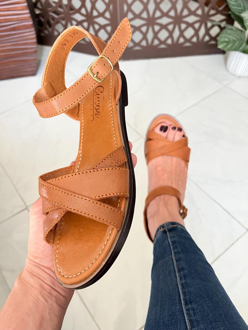 Pu&ograve; includere: Sandali in pelle marrone con un design a cinturino incrociato e una chiusura regolabile con fibbia. I sandali hanno un plantare imbottito e una suola in gomma.