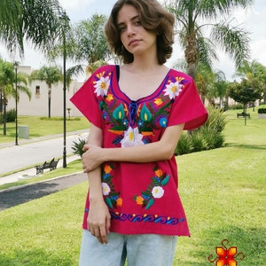 May include: A pink embroidered blouse with colorful floral designs. The blouse has short sleeves and a round neckline. The embroidery features flowers, leaves, and swirls in shades of blue, green, yellow, orange, and white.