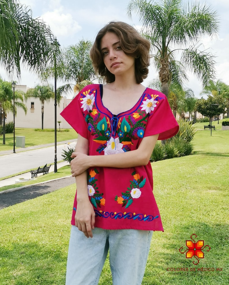May include: A pink embroidered blouse with colorful floral designs. The blouse has short sleeves and a round neckline. The embroidery features flowers, leaves, and swirls in shades of blue, green, yellow, orange, and white.