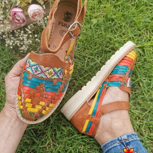 May include: Brown leather sandals with colorful woven straps. The sandals feature a buckle closure and a white sole. The design incorporates vibrant hues of red, yellow, blue, and green, creating a striking visual appeal. The sandals are displayed on a grassy surface.
