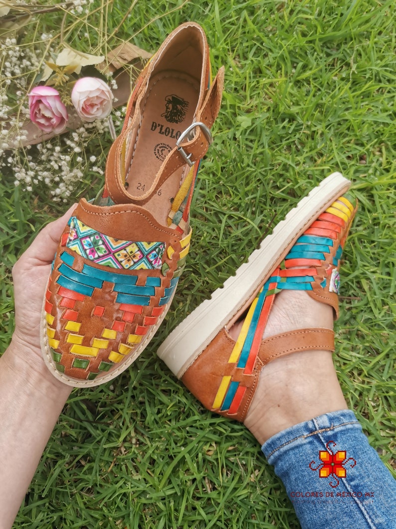 May include: Brown leather sandals with colorful woven straps. The sandals feature a buckle closure and a white sole. The design incorporates vibrant hues of red, yellow, blue, and green, creating a striking visual appeal. The sandals are displayed on a grassy surface.