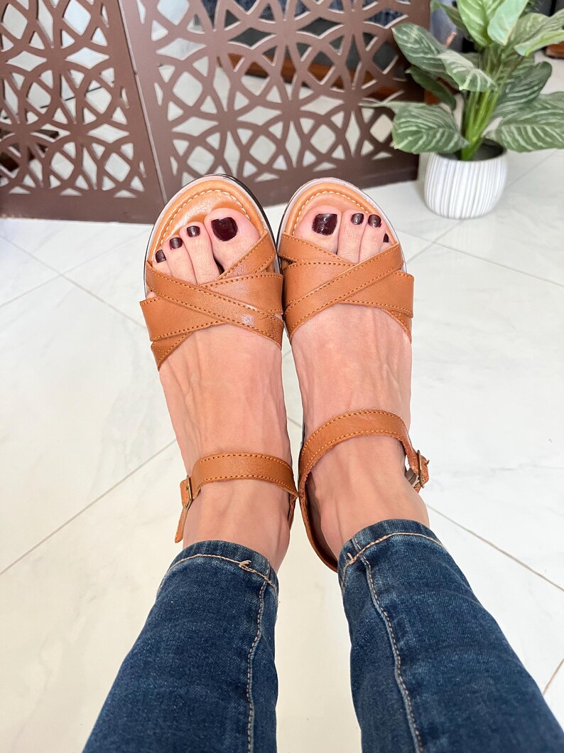 Pu&ograve; includere: Un paio di sandali in pelle marrone con cinturini incrociati e chiusure a fibbia. I sandali sono indossati con jeans blu.