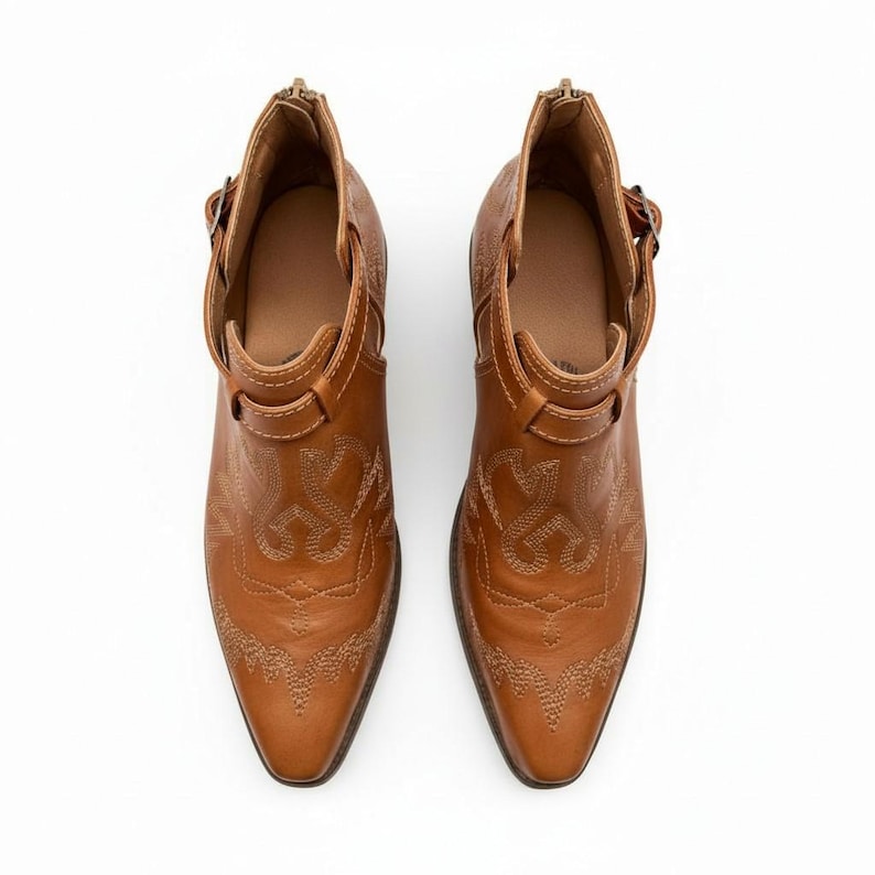 Pu&ograve; includere: Un paio di stivaletti alla caviglia in pelle marrone con un design di ispirazione western. Gli stivali presentano cuciture decorative, punte a punta e un piccolo tacco. Hanno una chiusura con cerniera sul retro e un dettaglio con fibbia laterale.
