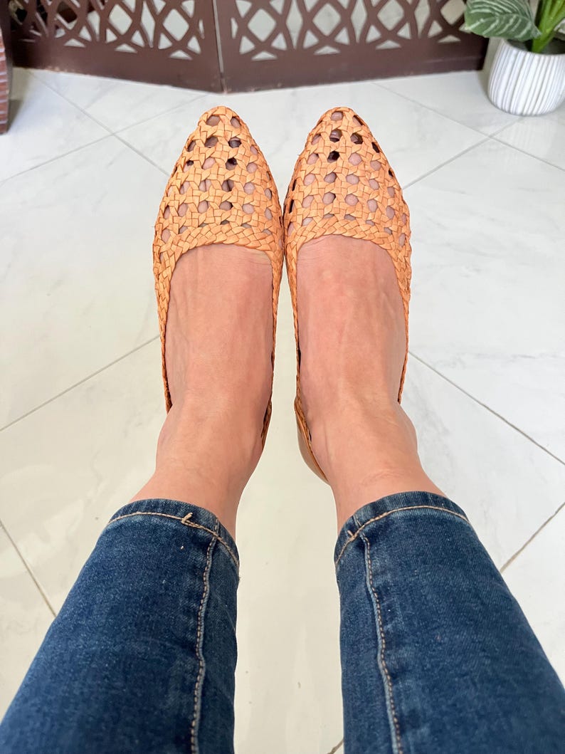 K&ouml;nnte beinhalten: Ein Paar geflochtene Lederschuhe in hellbrauner Farbe. Die Schuhe haben eine spitze Zehenpartie und ein geflochtenes Design. Die Schuhe werden von einer Person getragen, die eine blaue Jeans tr&auml;gt.