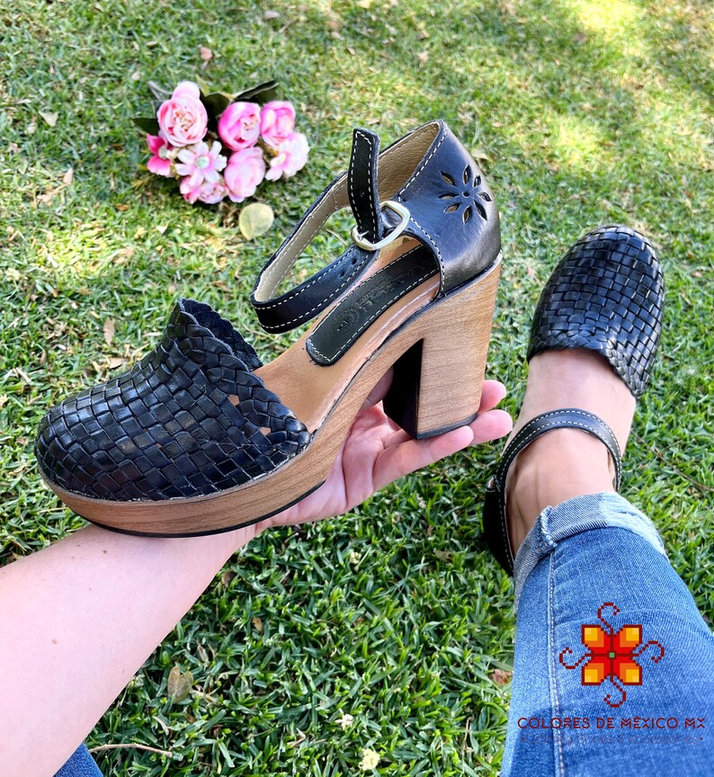 Op de afbeelding: Zwarte geweven leren platform sandalen met een dikke houten hak en een enkelband. De sandalen liggen op een groene grasmat.