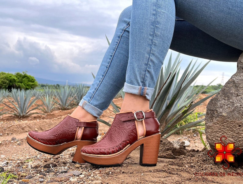 May include: A pair of brown leather heeled shoes with a cutout design and a wood platform sole. The shoes are worn by a person wearing blue jeans and sitting on a rock.