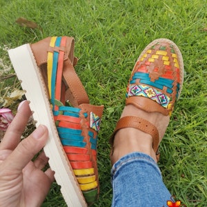 May include: Brown leather huaraches sandals with colorful woven straps in yellow, red, blue, and green. The sandals have a white sole and a strap around the ankle. The image shows the sandals on a grassy surface.