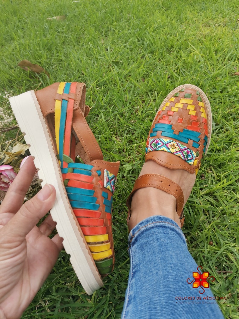 May include: Brown leather huaraches sandals with colorful woven straps in yellow, red, blue, and green. The sandals have a white sole and a strap around the ankle. The image shows the sandals on a grassy surface.
