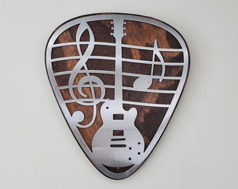 May include: A wooden guitar pick with a metal inlay of a guitar and musical notes. The pick is brown with a dark wood grain.