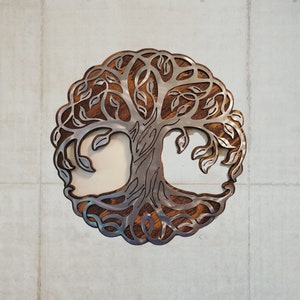 K&ouml;nnte beinhalten: Metall-Wandkunst mit dem Lebensbaum-Motiv. Das Design zeigt eine detaillierte Baum-Silhouette mit filigranen Blatt- und Astmustern. Der Baum ist vor einem h&ouml;lzernen Hintergrund platziert und von einem kreisf&ouml;rmigen Rahmen umgeben. Das Kunstwerk ist an einer Betonwand befestigt.