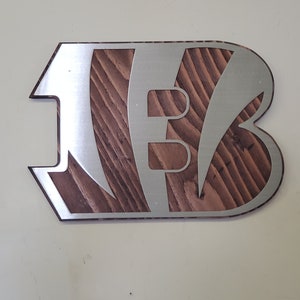 A steel angular emblem with diagonal stripe cutouts over rustic stained wood, shown on a light background.