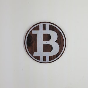 May include: A round wooden sign with a silver Bitcoin symbol on it. The sign is dark brown with a light brown wood grain.