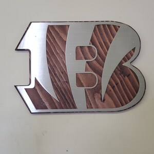 Angled view of a bold angular steel emblem with diagonal stripe cutouts over rustic stained wood, on a pale background.