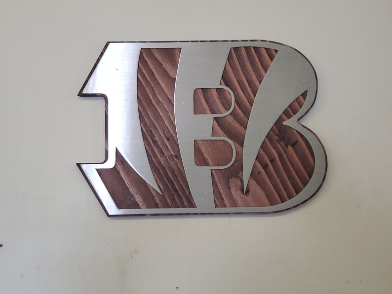Angled view of a bold angular steel emblem with diagonal stripe cutouts over rustic stained wood, on a pale background.