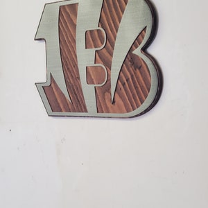 A steel angular emblem with diagonal stripe details over rustic stained wood, shown on a light gray backdrop.