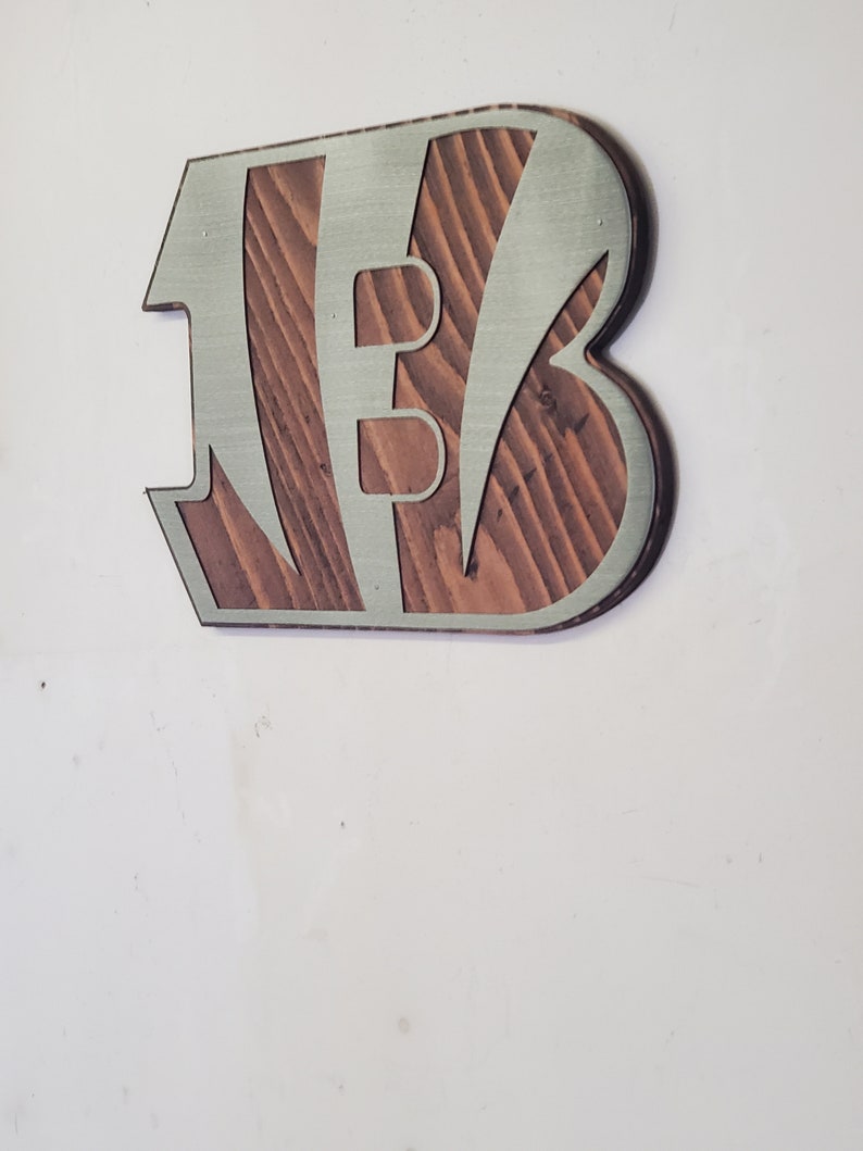 A steel angular emblem with diagonal stripe details over rustic stained wood, shown on a light gray backdrop.