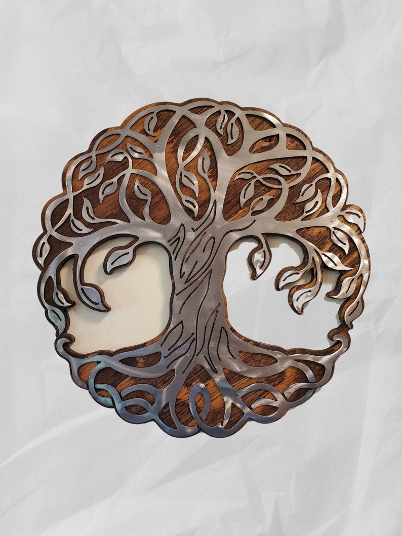 K&ouml;nnte beinhalten: Runde Wanddekoration mit einem Lebensbaum-Motiv. Der Baum ist aus silberfarbenem Metall gefertigt und vor einem Holzgrund platziert. Das Design umfasst detaillierte &Auml;ste, Bl&auml;tter und Wurzeln, wodurch ein symboltr&auml;chtiges Dekor entsteht.