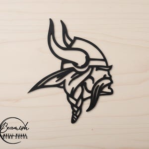 May include: Black metal wall art featuring the profile of a Viking with a horned helmet and braided beard. The artwork is set against a light wood grain background. The logo for Beamish Metal Works Home Decor is in the lower left corner.