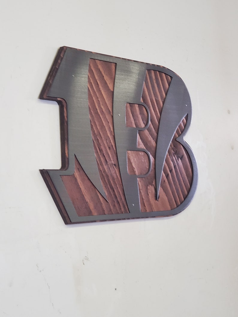 A steel angular emblem with diagonal stripe cutouts over rustic stained wood, angled on a light wall with a darker steel finish.