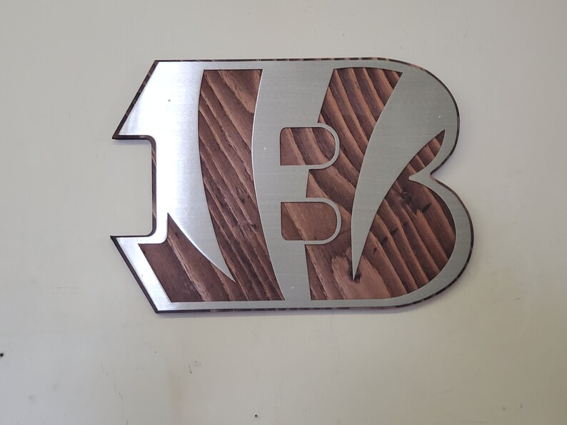 A steel angular emblem with diagonal stripe cutouts over rustic stained wood, shown on a light background.