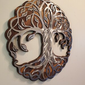 K&ouml;nnte beinhalten: Runde Wanddekoration mit einem Baum des Lebens-Design. Der Baum besteht aus silberfarbenem Metall und ist vor einem dunkelbraunen Holzgrund platziert. Der &auml;u&szlig;ere Rand des Kunstwerks ist ebenfalls dunkelbraun.