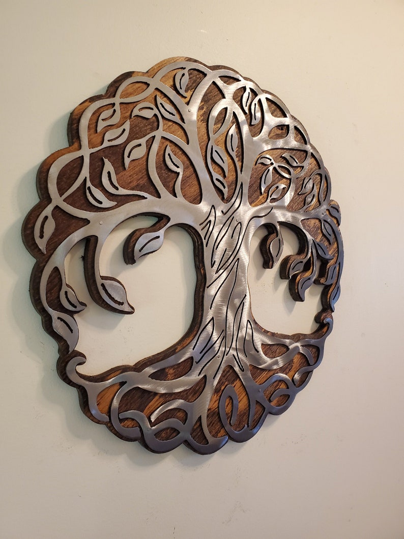 K&ouml;nnte beinhalten: Runde Wanddekoration mit einem Baum des Lebens-Design. Der Baum besteht aus silberfarbenem Metall und ist vor einem dunkelbraunen Holzgrund platziert. Der &auml;u&szlig;ere Rand des Kunstwerks ist ebenfalls dunkelbraun.