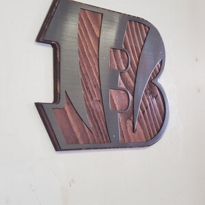 A steel angular emblem with diagonal stripe cutouts over rustic stained wood, angled on a light wall with a darker steel finish.