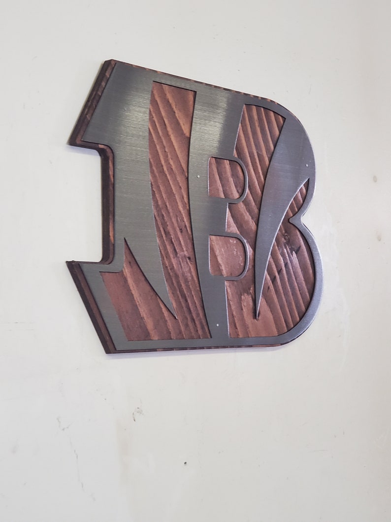 A steel angular emblem with diagonal stripe cutouts over rustic stained wood, angled on a light wall with a darker steel finish.