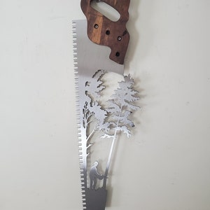May include: A metal saw blade with a wooden handle. The blade is cut out in the shape of a tree and a person cutting down the tree.