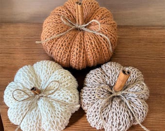 May include: Three handmade knitted pumpkins in shades of orange, cream, and beige. Each pumpkin features a cinnamon stick stem and is tied with twine. The pumpkins are arranged on a wooden surface, perfect for fall decor.