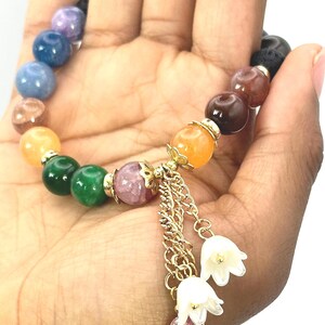 May include: A beaded bracelet with a variety of colorful gemstones, including blue, green, orange, and purple. The bracelet has a gold chain with white flower charms and small beads. The bracelet is held in a hand.