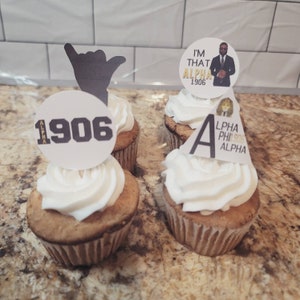 May include: Four cupcakes decorated with white frosting and various toppers. One cupcake has a black silhouette of a hand, another has a white circle with the text "1906" and "ALPHA", another has a white circle with a man in a suit and the text "I'M THAT ALPHA 1906", and the last cupcake has a gold triangle with the text "ALPHA PHI 1906 ALPHA".