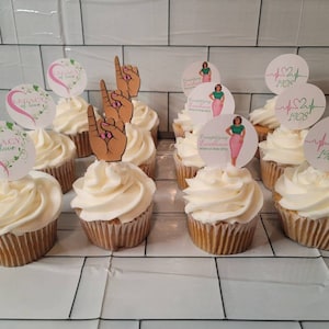 May include: A dozen white cupcakes with pink and green frosting. The cupcakes are decorated with various toppers, including a heart with the text "Legacy of Love", a hand gesture, and a heart with the text "Celebrating Excellence 1908".